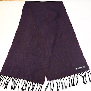 May include: A dark burgundy scarf with black fringe. The scarf is made of a woven material and has a rectangular shape. A small black tag with white text is visible on the scarf. The scarf is a winter accessory.