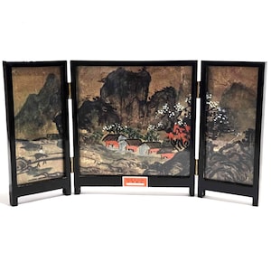 Può includere: Uno schermo in legno nero a tre pannelli con una scena di paesaggio dipinta. La scena presenta montagne, alberi e un villaggio con tetti rossi. Lo schermo ha un'etichetta che recita "Made in China".