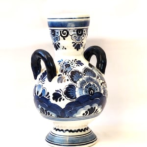 May include: A white ceramic vase with blue and white floral designs and black handles. The vase has a round body and a flared base.