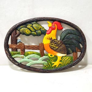 Könnte beinhalten: Gusseisen-Topfuntersetzer mit einem bunten Hahn mit rotem Kamm und Kehllappen, gelbem Körper, braunen und schwarzen Federn und grünem Schwanz. Der Hahn steht auf einer grünen Grasfläche mit einem braunen Zaun und einem grünen Baum im Hintergrund.