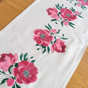 Puede incluir: Camino de mesa blanco con un estampado floral. El camino presenta grandes flores rosas estilizadas con centros rojo oscuro y hojas verdes. Las flores están dispuestas en un patrón repetitivo a lo largo, creando un acento decorativo para una mesa de comedor.