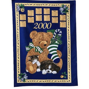 Puede incluir: Un paño de cocina azul con un borde amarillo presenta un oso de peluche marrón que lleva una media a rayas verdes y blancas, un gatito marrón y blanco durmiendo en el regazo del oso y un cachorro marrón y blanco durmiendo a los pies del oso. El paño también presenta un calendario para el año 2000.
