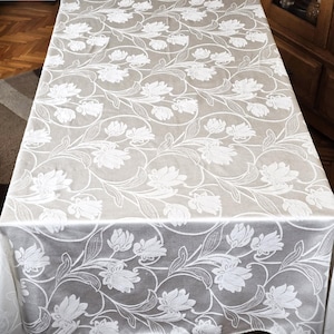 Peut inclure: Nappe rectangulaire blanc cassé avec un motif floral répété. Les fleurs et les feuilles blanches créent un motif texturé. La nappe est drapée sur une table, montrant toute sa surface. Le tissu a un éclat subtil.