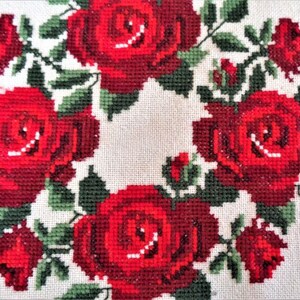May include: A white fabric with a red embroidered wreath of roses. The roses are in a circular pattern with green leaves.