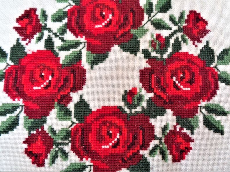 May include: A white fabric with a red embroidered wreath of roses. The roses are in a circular pattern with green leaves.