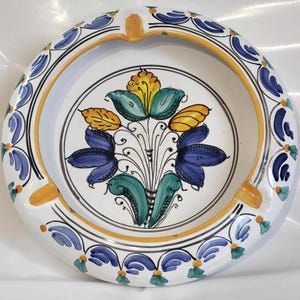 May include: A white ceramic ashtray with a blue and yellow floral design. The ashtray has a round shape and a wide rim.