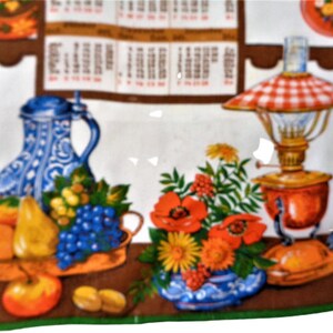 Op de afbeelding: Een vintage keukendoek met een kleurrijk ontwerp met een blauw-witte kan, fruit, bloemen en een rood-wit geblokte olielamp.