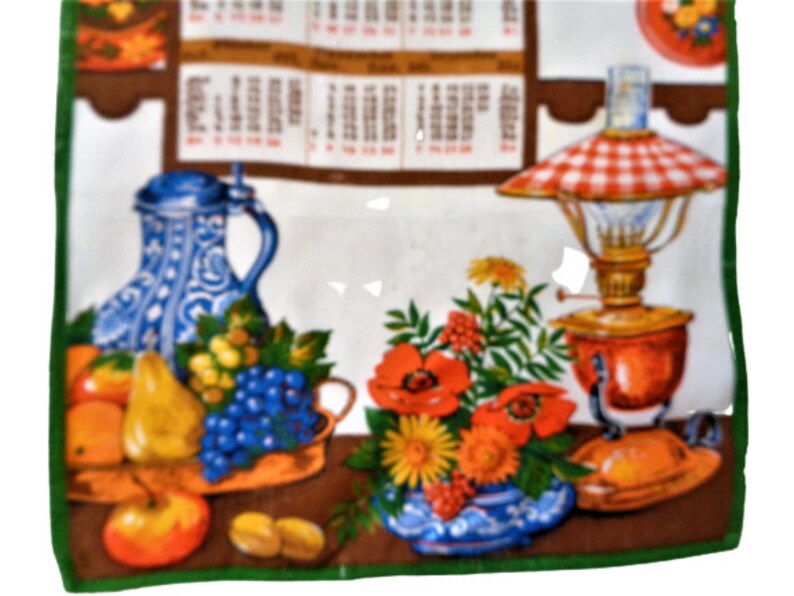 Op de afbeelding: Een vintage keukendoek met een kleurrijk ontwerp met een blauw-witte kan, fruit, bloemen en een rood-wit geblokte olielamp.
