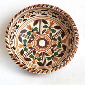 Może przedstawiać: Biała ceramiczna miska z brązowym, pomarańczowym i zielonym wzorem kwiatowym. Miska ma podwyższony rant z brązowym i pomarańczowym wzorem sznurkowym.