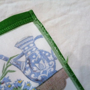 Op de afbeelding: Een witte stoffen servet met een groene rand. De servet heeft een blauwe en witte kan met bloemenpatronen en groene bladeren. De kan staat op een bruin oppervlak.