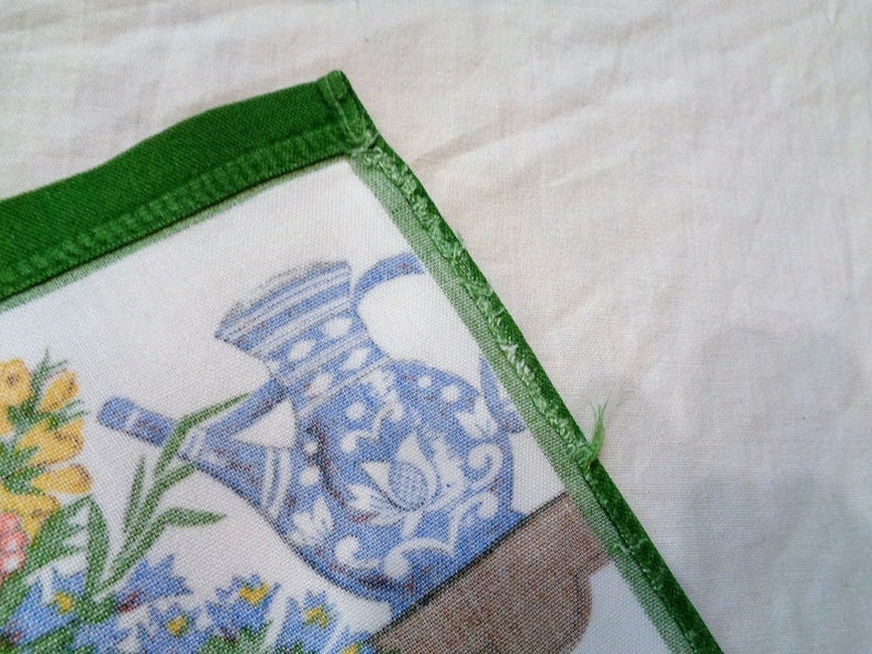 Op de afbeelding: Een witte stoffen servet met een groene rand. De servet heeft een blauwe en witte kan met bloemenpatronen en groene bladeren. De kan staat op een bruin oppervlak.