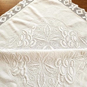 May include: Ivory-colored vintage linen tablecloth with intricate white embroidered grape and leaf design. The tablecloth features a delicate lace trim along the edges, adding a touch of elegance. The embroidery is detailed and creates a textured surface.