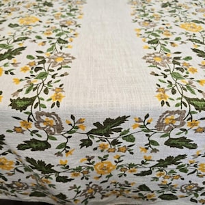 May include: A white linen tablecloth featuring a repeating floral pattern with yellow blossoms, green leaves, and grey-brown flower centers arranged in vertical bands along the sides, leaving a plain white central strip of fabric.