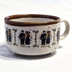 Könnte beinhalten: Eine braune und weiße Keramiktasse mit braunem Rand und Henkel. Die Tasse hat ein Design mit Paaren in traditioneller niederländischer Kleidung mit dem Wort "Zeeland" auf der Tasse.