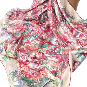 Puede incluir: Un pañuelo de seda color crema con un vibrante diseño floral y de volutas en tonos rojos, rosas, verdes y morados. El pañuelo tiene un borde rojo y la marca "yanensis" es visible en la esquina.