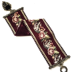 May include: A burgundy velvet ribbon with a floral tapestry design. The ribbon is trimmed with gold lace and ornate metal hardware at each end. The floral pattern features roses and other flowers in shades of pink, cream, and green.