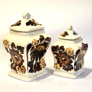 Op de afbeelding: Twee keramische potten met deksels, beide versierd met bruine en gele bloemenpatronen. De potten zijn vierkant met een licht afgeronde boven- en onderkant.