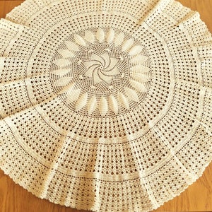 Könnte beinhalten: Eine große, runde, gehäkelte Tischdecke in Cremefarbe mit einem detaillierten, komplizierten Muster. Die Tischdecke hat einen gewellten Rand.