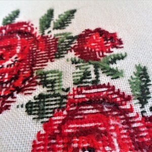 May include: Close-up of a white fabric with red and green embroidered roses. The roses are stitched with a cross-stitch pattern.