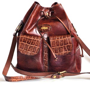 Könnte beinhalten: Eine braune Ledereimer-Tasche mit Kordelzugverschluss und einem langen, verstellbaren Trageriemen. Die Tasche hat zwei Fronttaschen mit einem Krokodilprägemuster und einem goldfarbenen Logo.