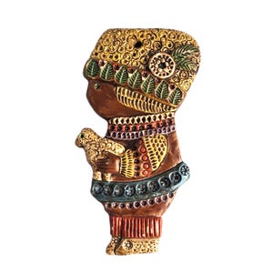 Può includere: Statuetta decorativa di una persona che tiene un piccolo animale. La statuetta è principalmente marrone con accenti dorati, verdi e blu. La persona indossa un copricapo e abiti dettagliati e l'animale è marrone chiaro.