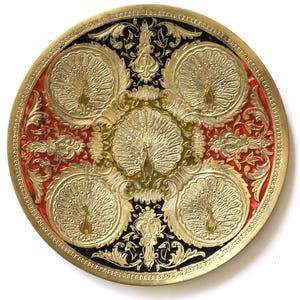 Op de afbeelding: Omsierd, rond decoratief bord met een gouden pauwenontwerp. Het bord heeft een rode en zwarte achtergrond met gouden filigraan en een parelrand. Het pauwenontwerp wordt vijf keer herhaald.