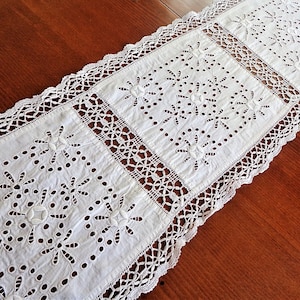 May include: White crocheted table runner with a delicate floral pattern. The runner has a lace border and is made of a lightweight fabric.