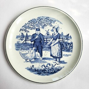 Puede incluir: Plato de cerámica azul y blanco con una escena holandesa con un hombre y una mujer con ropa tradicional, un molino de viento y gansos. El plato tiene un borde negro.