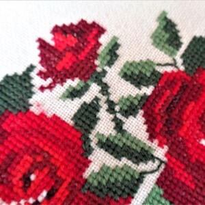 May include: Close-up of a white fabric with red and green embroidered roses. The roses are stitched in a cross-stitch pattern.