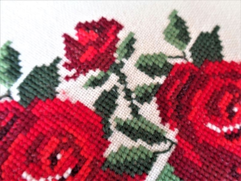 May include: Close-up of a white fabric with red and green embroidered roses. The roses are stitched in a cross-stitch pattern.