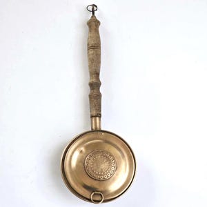 May include: A vintage brass pan with a wooden handle and a decorative design on the bottom. The pan has a small ring on the side.