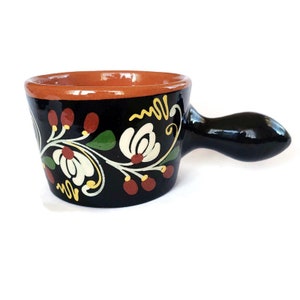 May include: A small black ceramic pot with a brown rim and a black handle. The pot is decorated with a white floral design with yellow and green accents.