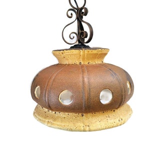 Puede incluir: Una lámpara colgante de cerámica marrón y amarilla con una cadena de suspensión de hierro forjado. La lámpara tiene un diseño redondeado en forma de cúpula con múltiples recortes circulares.