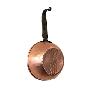 May include: A copper saucepan with a hammered bottom and a dark metal handle. The handle curves upward and has a small loop at the end. The pan is round and has a shiny, reflective surface.