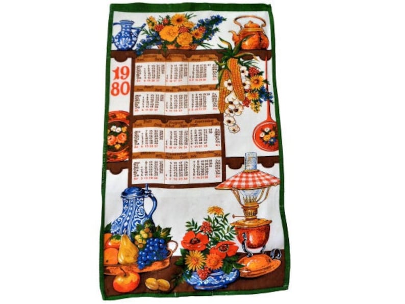 Op de afbeelding: Een vintage keukendoek met een groene rand en een witte achtergrond. De handdoek heeft een kalender voor het jaar 1980, een stilleven met fruit, bloemen en keukengerei, en de tekst "1980".