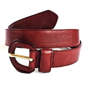 Op de afbeelding: Een dieprode riem met een gestructureerd hagedis-huidpatroon over de gehele band en de ovale gesp. De riem is voorzien van een bijpassende lus en een gepolijste goudkleurige metalen gesptong die door het frame van de gesp steekt.