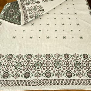 Könnte beinhalten: Cremefarbene Tischdecke mit detailliertem floralem Rand in Braun-, Grün- und Burgundertönen. Das Design umfasst sich wiederholende Blumen- und geometrische Muster. Der gefaltete Rand zeigt eine grün-braun gestreifte Unterseite.