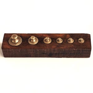 Op de afbeelding: Een vintage set messing gewichten in een donkerhouten houder. De gewichten zijn in een rij gerangschikt, met verschillende maten en markeringen. De houten basis heeft een rijke, donkerbruine kleur en de messing gewichten hebben een warme, gouden tint.
