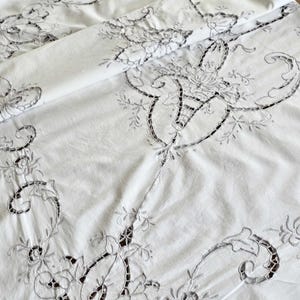 Può includere: Tovaglia bianca con ricami floreali e dettagli traforati. Il design presenta fiori delicati, foglie e motivi a volute. Il tessuto è piegato, mostrando l'artigianato dettagliato e la consistenza del tessuto vintage.