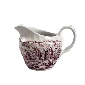 Può includere: Un creamer in ceramica bianca con un design floreale bordeaux che rappresenta un castello. Il creamer ha un manico e un beccuccio.
