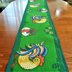 Op de afbeelding: Groene tafelrunner met een herhalend patroon van gele kippen, blauwe en roze veren en rode en witte eieren met strikken. De runner heeft een groene rand met een wit en zwart geruit patroon.