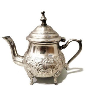 Peut inclure: Une théière en argent avec un design orné et un couvercle à charnière. La théière a trois pieds et un bec.