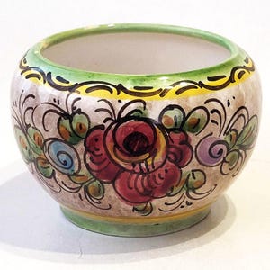 Può includere: Un piccolo vaso rotondo in ceramica con fondo bianco e design floreale dipinto a mano. Il vaso presenta una grande rosa rossa con accenti gialli e verdi, insieme a fiori blu e viola più piccoli.