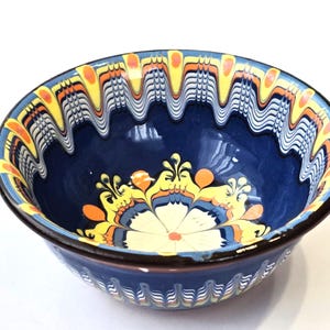 Puede incluir: Un cuenco de cerámica azul con un borde decorativo con un patrón repetitivo de líneas onduladas amarillas, naranjas y blancas. El interior del cuenco tiene un diseño floral en amarillo, naranja y blanco. El borde es marrón oscuro.