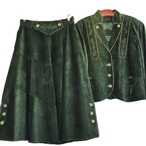 May include: A dark green suede skirt and jacket set. The skirt is a-line with decorative buttons and a detailed waistband. The jacket has gold trim, decorative embroidery, and matching buttons. The set is displayed on a white background.