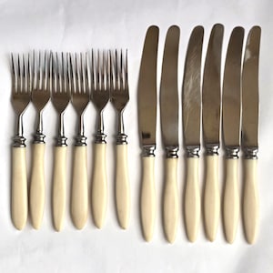 Peut inclure: Un ensemble de couverts anciens, comprenant des fourchettes et des couteaux. Les fourchettes ont quatre dents et les couteaux ont une lame incurvée. Les deux ont des manches crème et des détails en métal argenté.