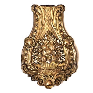 Peut inclure: Une plaque murale de couleur dorée présentant un relief central représentant un panier rempli de fleurs. La plaque possède un cadre orné et symétrique avec des volutes, des motifs de feuilles et une texture de fond en losanges.