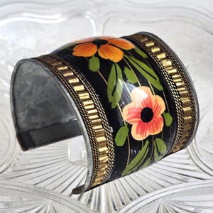 Può includere: Un ampio bracciale a polsino in metallo con finitura smaltata nera. Il bracciale è decorato con fiori arancioni e rossi dipinti a mano con foglie verdi. Le finiture in metallo color oro accentuano i bordi del bracciale, aggiungendo un tocco di eleganza al design floreale.