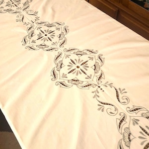 May include: White tablecloth with a delicate embroidered design featuring intricate floral patterns and geometric shapes. The embroidery is done in a silver thread, creating a subtle shimmer.