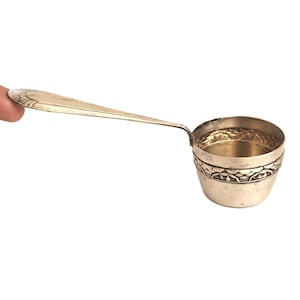 Könnte beinhalten: Eine silberne Schöpfkelle mit langem Griff und einer kleinen, runden Schüssel. Die Schüssel hat ein dekoratives Band um den oberen Rand mit einem geätzten Muster. Der Griff wird vom Finger einer Person gehalten.